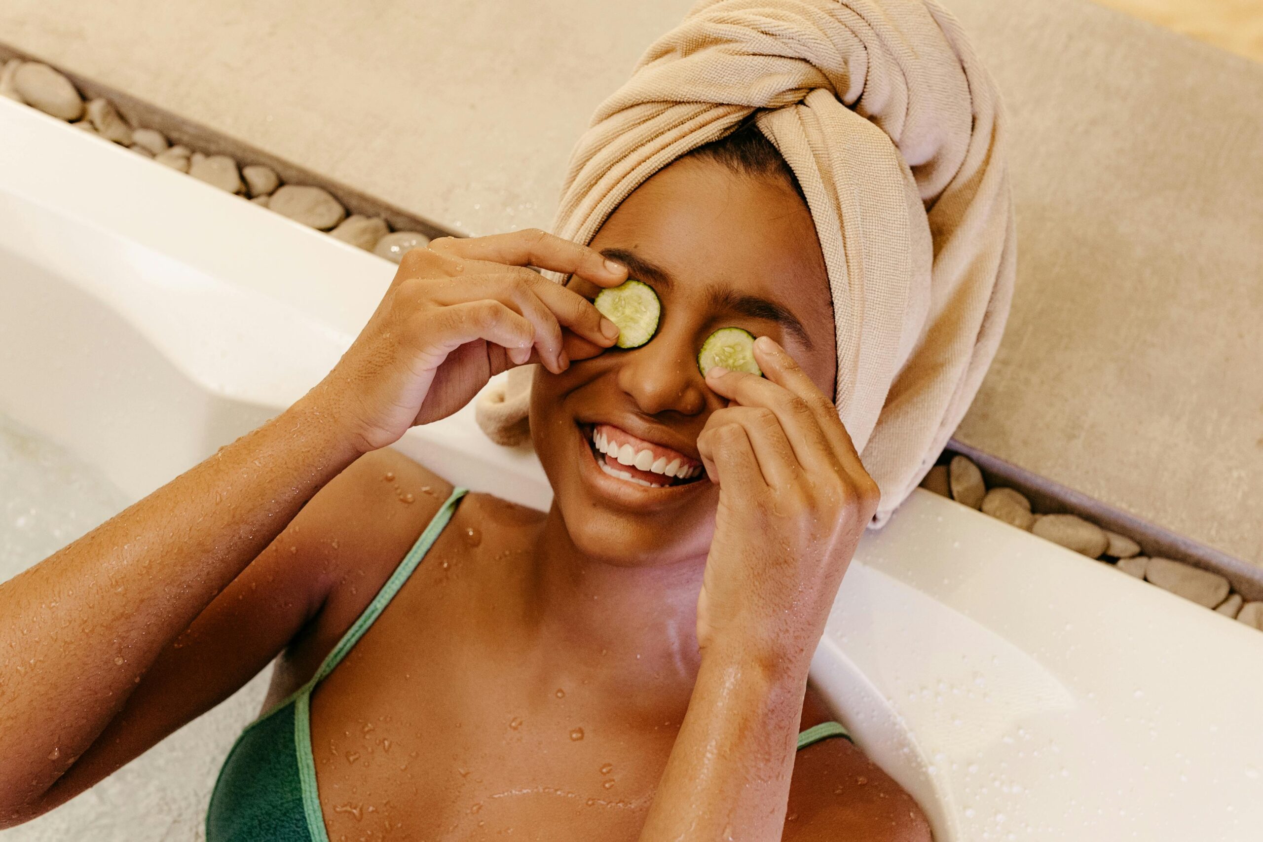 A woman relaxing in a jacuzzi at home with a towel wrapped around her hair and cucumber slices on her eyes, creating a spa experience to save money on her beauty routine