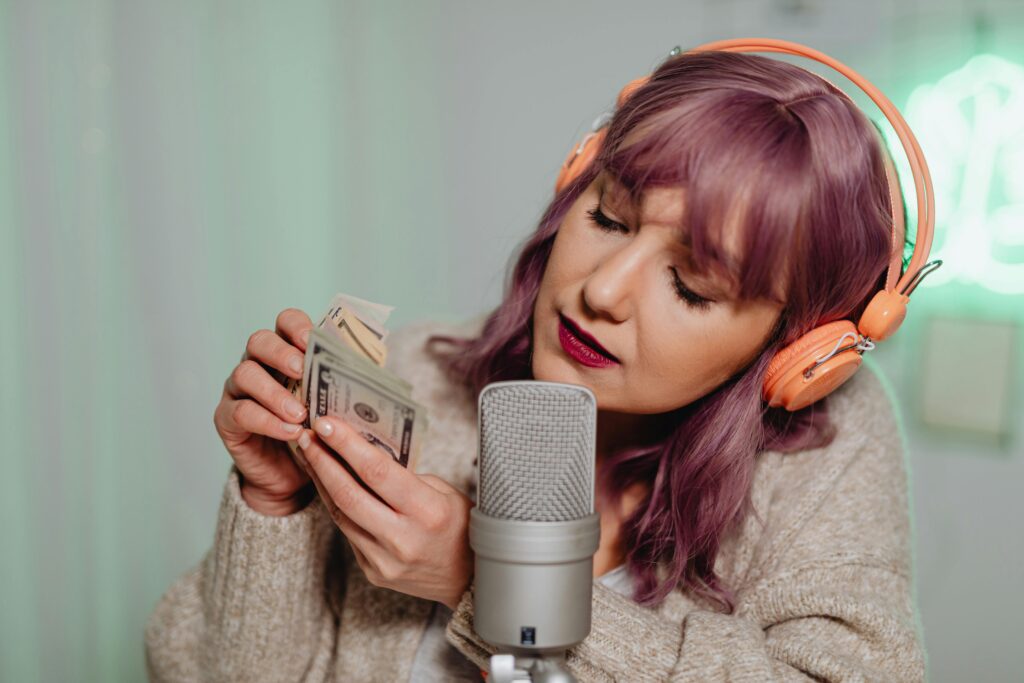 Woman holding cash in her hands, wearing headphones, sitting in front of a podcast mic, showcasing "How To Turn Your Creativity Into Passive/Royalty Income Online."